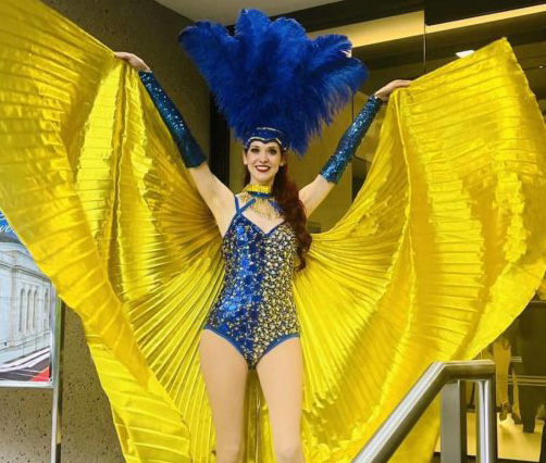 Costumed female performer raising a silk cape above her head in front of the B2B entrance.