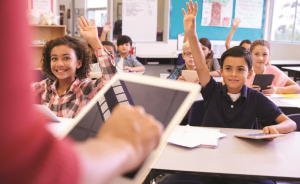 Smiling young children in a classroom raise their hand. A teacher's hand using a tablet is in the foreground.