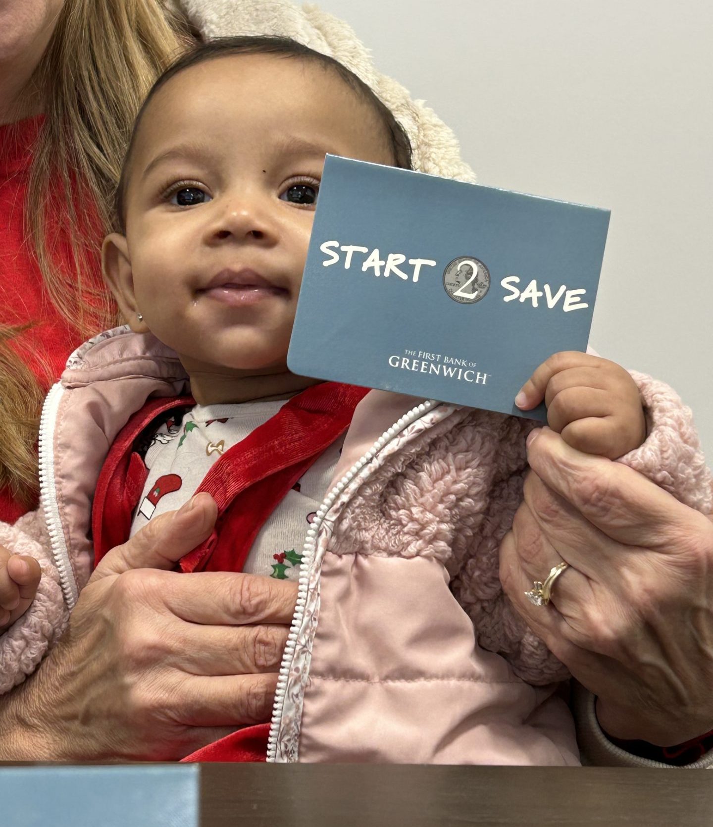An image of a female, Black toddler sitting on someone's lap holding a Start2Save book.