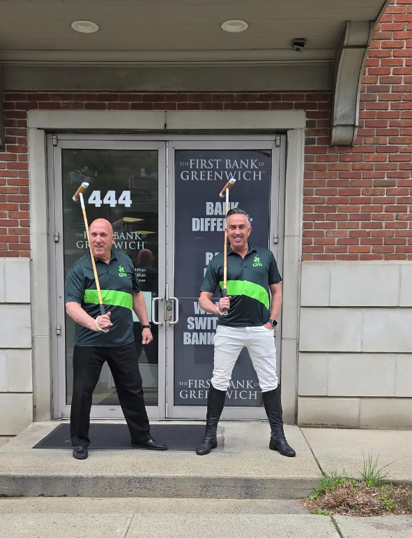 Bank President, Frank J. Gaudio, and Greenwich Polo Academy founder, Christian Perry, stand in from of the Bank's Cos Cob branch smiling while wearing GPA jerseys and holding polo mallets.