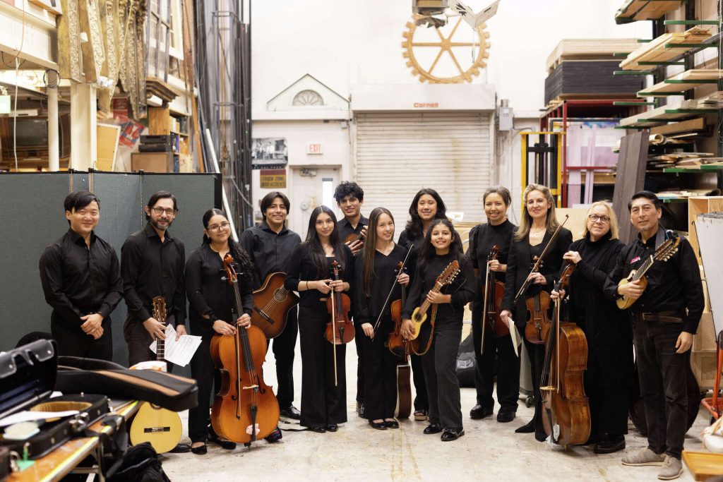 Lou Chen, CEO of INTEMPO, is standing in what appears to be a warehouse on the left-hand side of a group of members of Orchestra Lumos and their instruments.