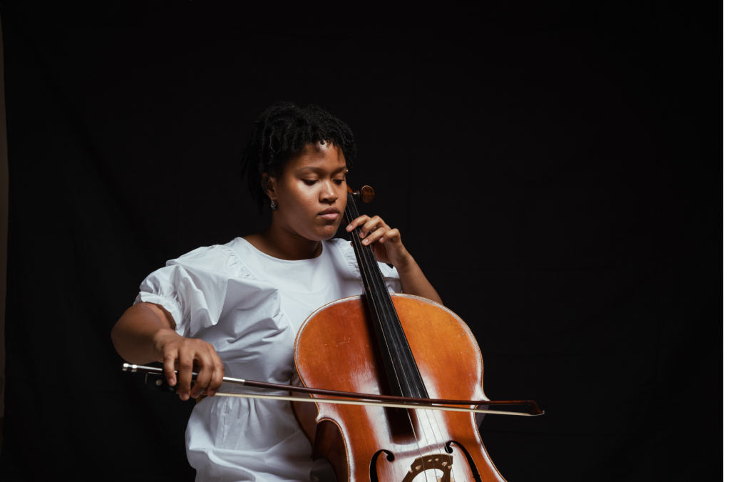 Internationally acclaimed cellist and composer Sujari Britt sits playing her cello, wearing a blue dress against a black background.