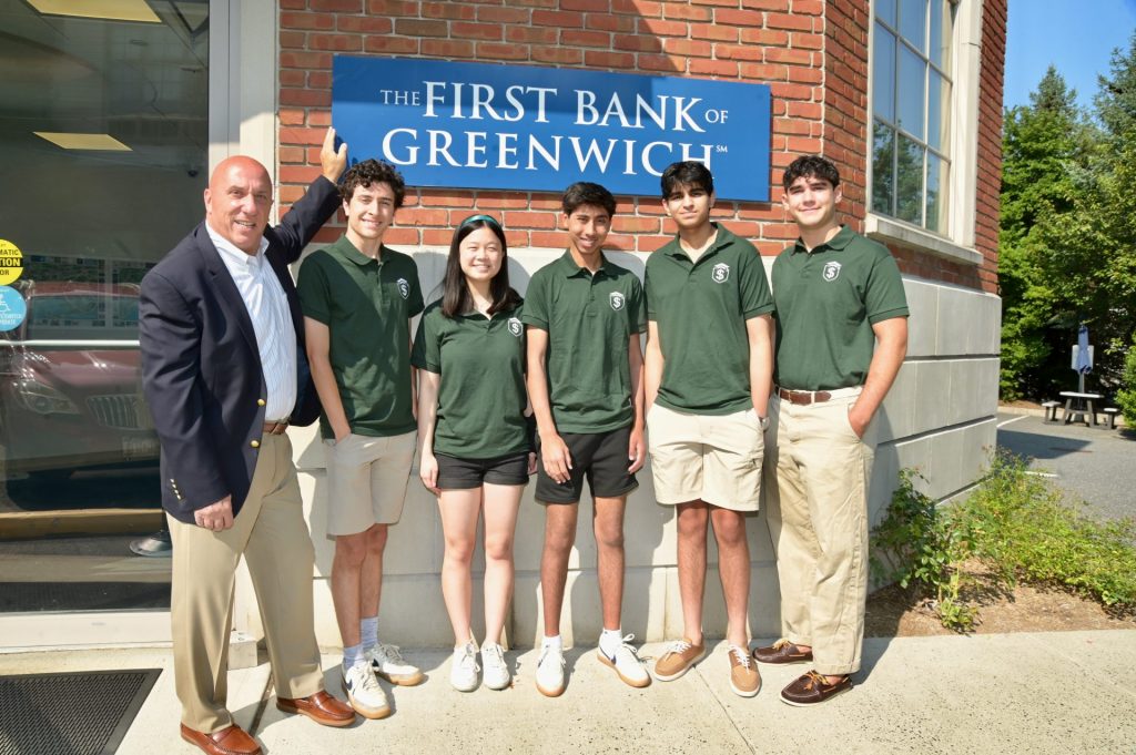 First Bank of Greenwich President & CEO stands outside in front of the Bank's sign with Sylvester Pecora, founder of the non-profit "Dollars to Scholars", and 4 other members of the Dollars for Scholars organization, a young Asian girl and 3 young men.