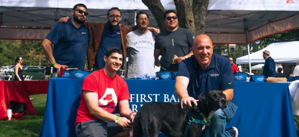 Frank J. Gaudio, the Bank's President and CEO, is pictured kneeling with a black Labrador Retriever and a member of the Adopt-A-Dog staff. 4 staff members from the Bank stand behind a table in the background.